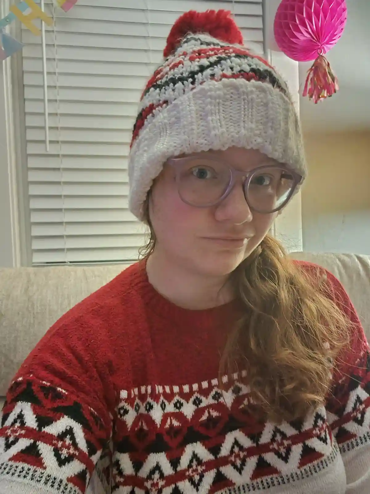 A person wearing a red and white patterned knit hat with a red pom-pom and a matching red holiday sweater sits on a couch…