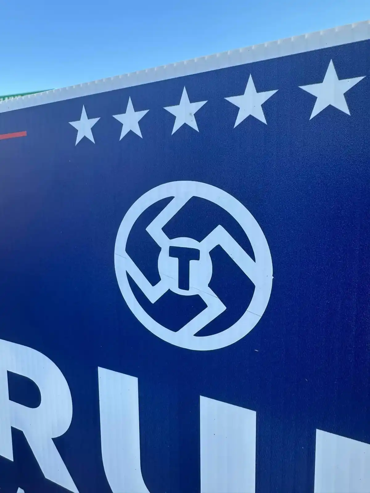 A close-up of a blue political campaign sign with five white stars along the top, a circular white logo containing a…