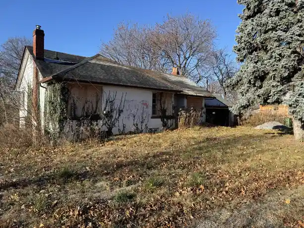 An abandoned, dilapidated house with boarded-up windows, overgrown ivy, and a neglected yard under a clear blue sky. The…