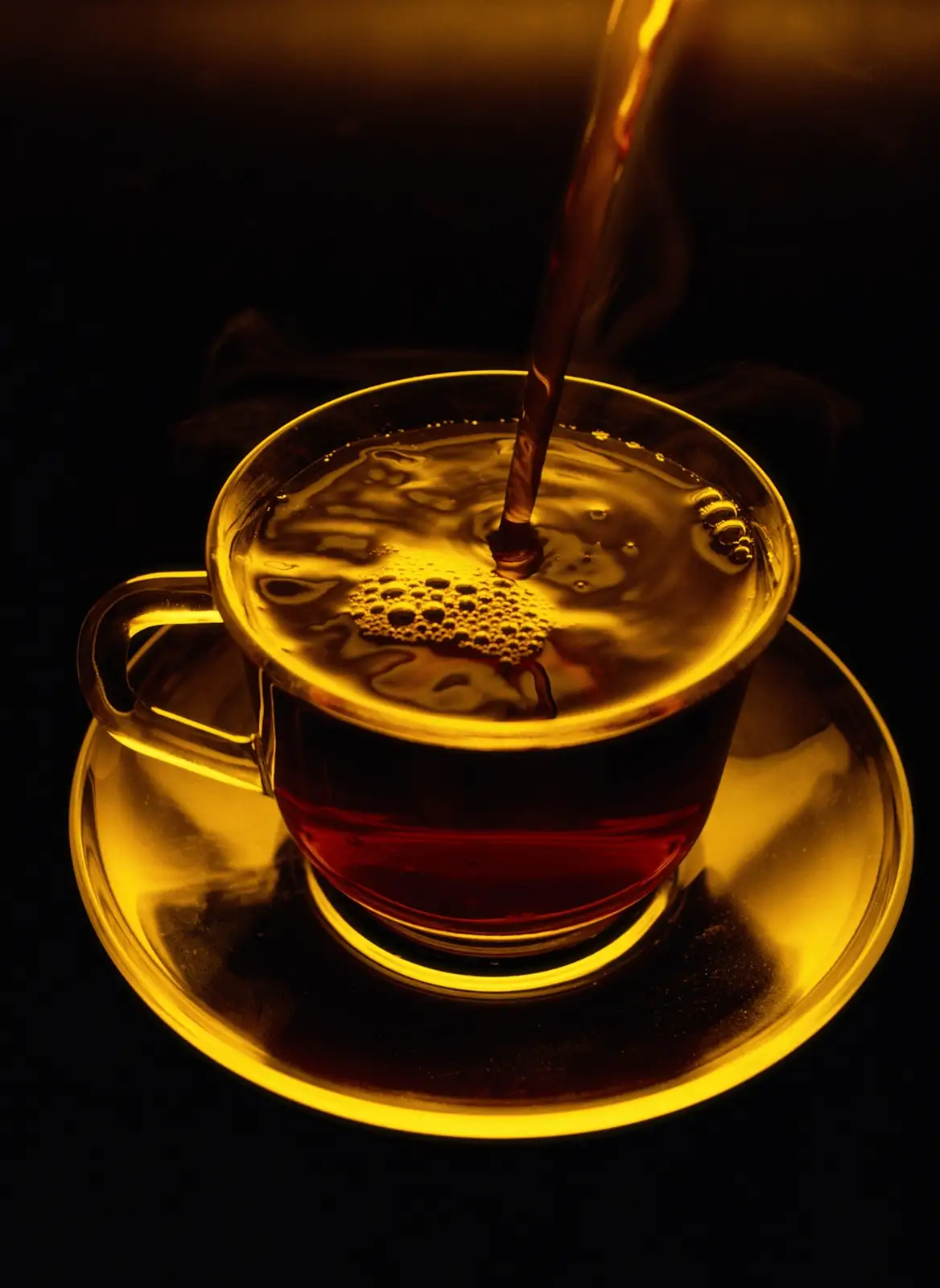 A close-up shot shows a dark amber liquid, presumably hot tea, being poured into a clear glass cup resting on a matching…