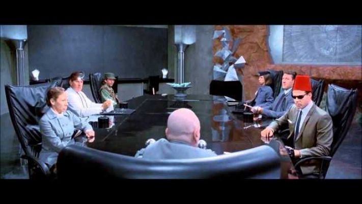 A still from a movie showing a tense meeting around a large black conference table. Several people are seated, including a…