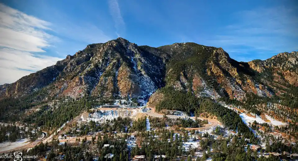 A scenic aerial photo of a snow-dusted mountain range with pine forests, winding roads, and a building complex at the base…