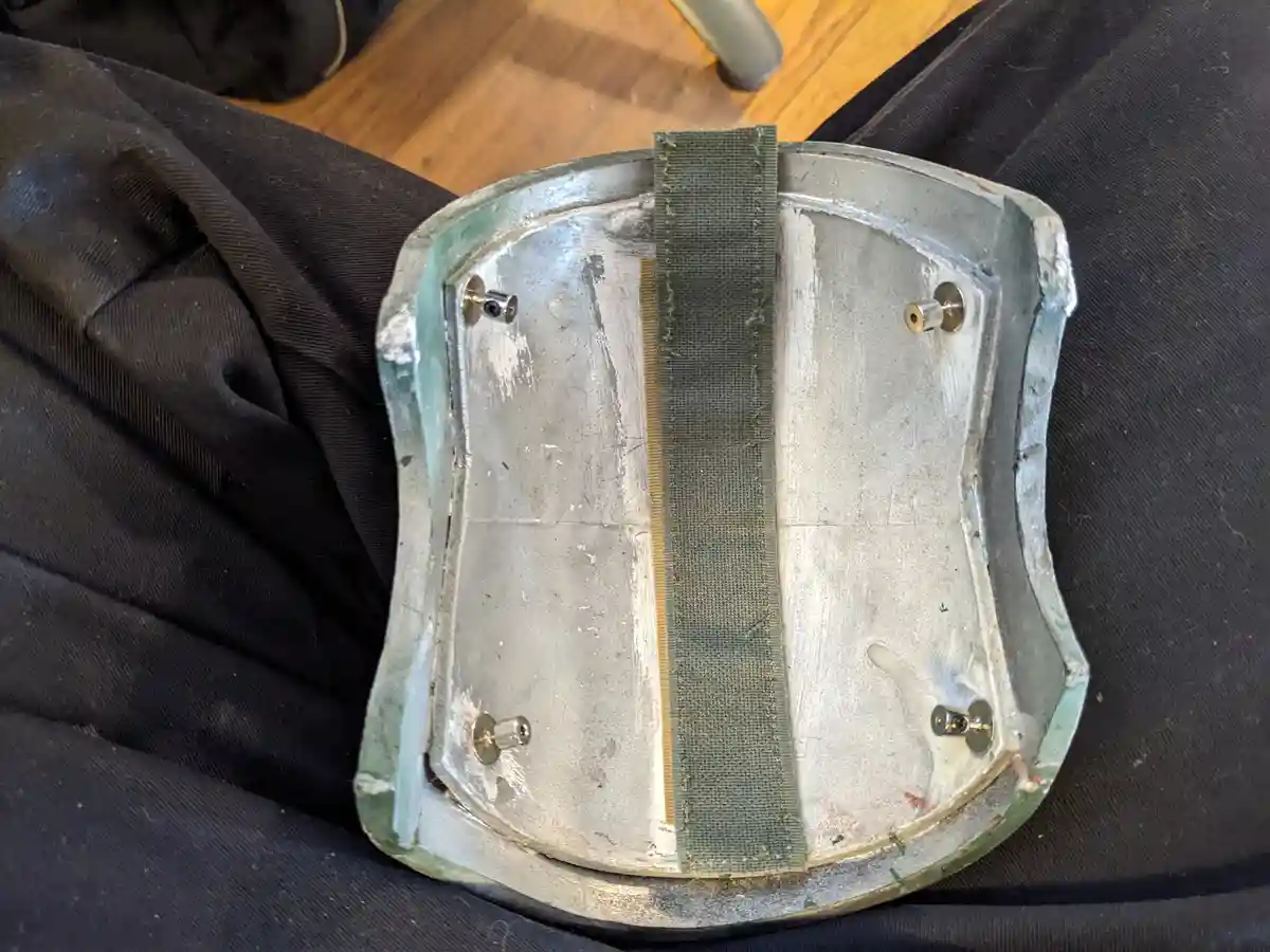 A close-up photo shows the underside of a homemade, silver-colored metal shield with four rivets and a green fabric strap…