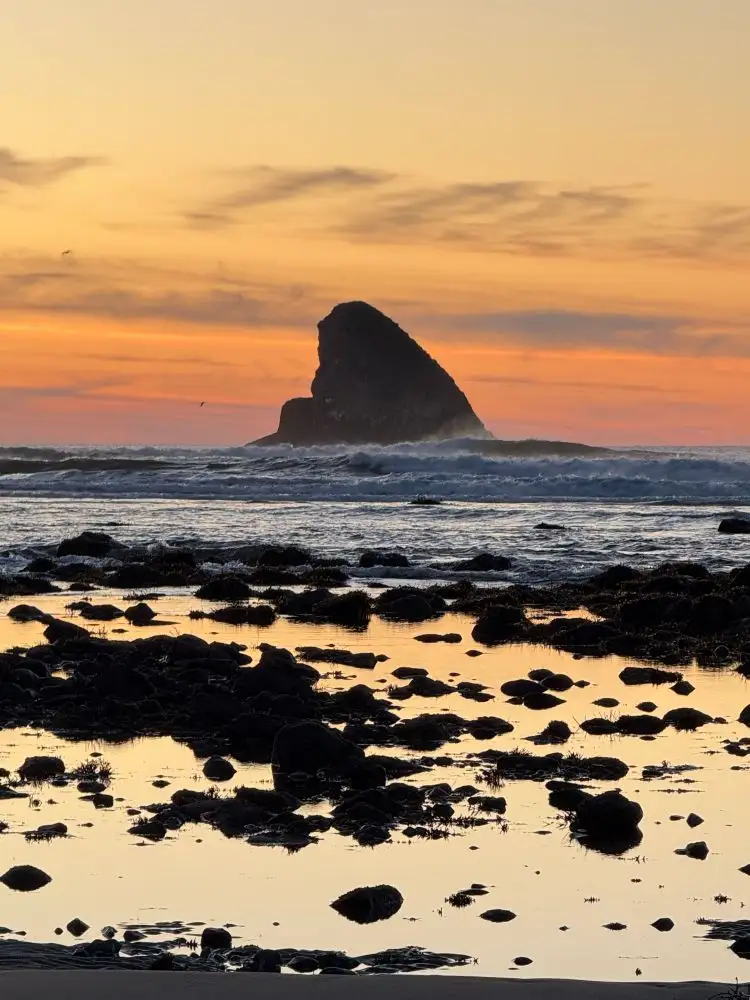 A serene sunset photograph of a large sea stack silhouetted against an orange and yellow sky, with waves crashing nearby and…