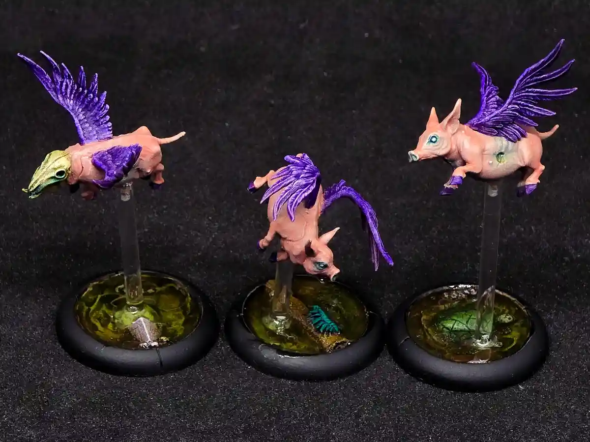 Three painted miniature pig-like creatures with purple wings are mounted on clear rods atop black circular bases against a…