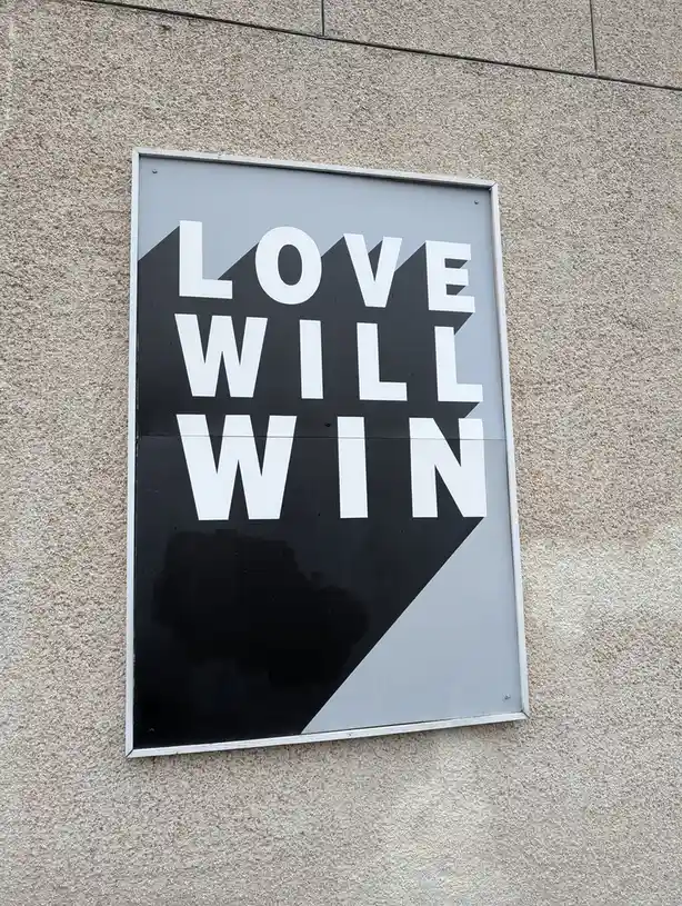 A black and white sign with bold white text reading 'LOVE WILL WIN' is mounted on a textured beige wall. The sign features a…