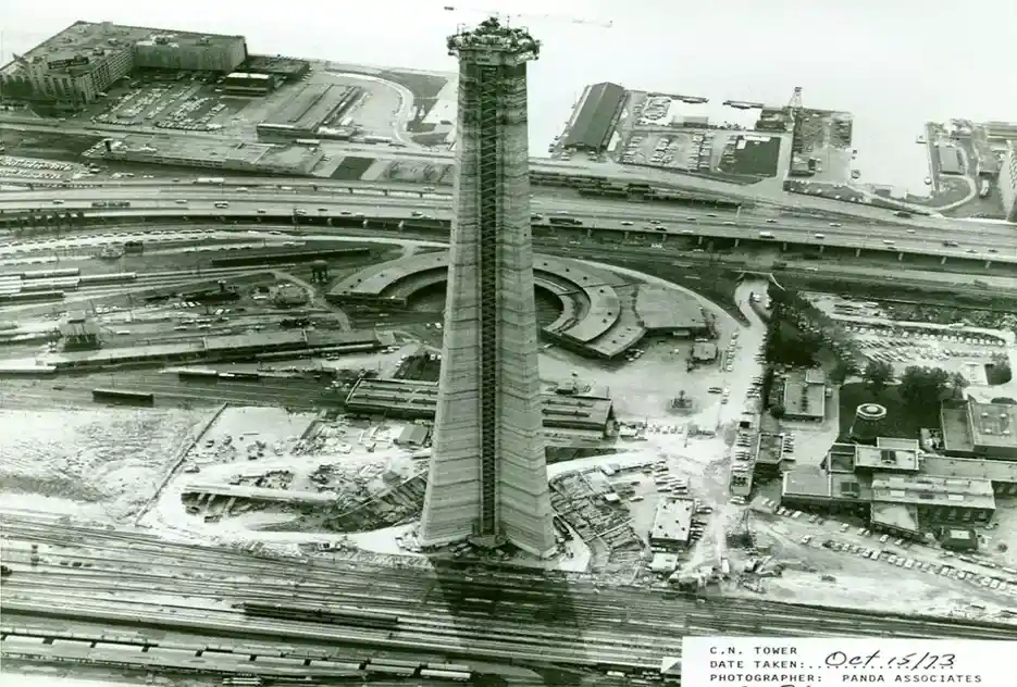 A black-and-white aerial photograph shows the partially constructed CN Tower in Toronto, surrounded by highways, railways…