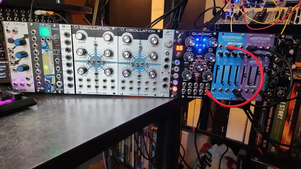 A photo of a modular synthesizer setup with multiple Eurorack modules like 'OSCILLATION', 'QUADNIC', and 'NOSTALGIA'…