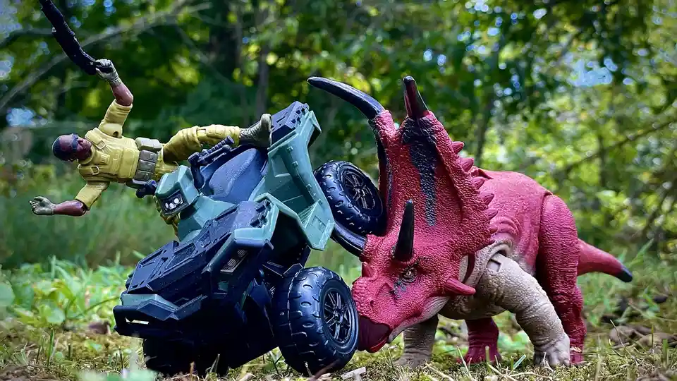 A toy triceratops with pink and beige coloring is shown pushing a green and black toy ATV, which has launched a yellow-clad…