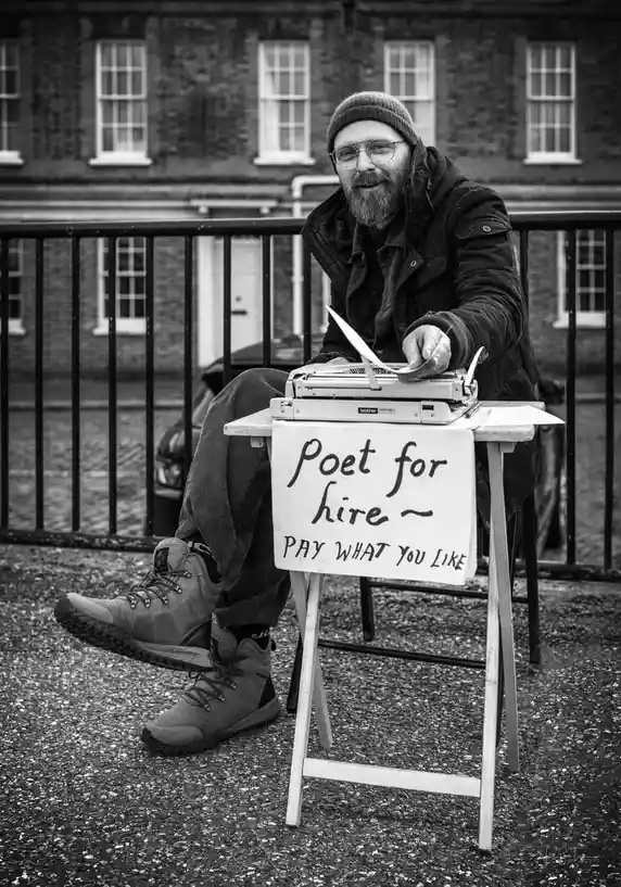 A black-and-white photo shows a bearded man with glasses and a beanie sitting outdoors on a chair, smiling at the camera. He…