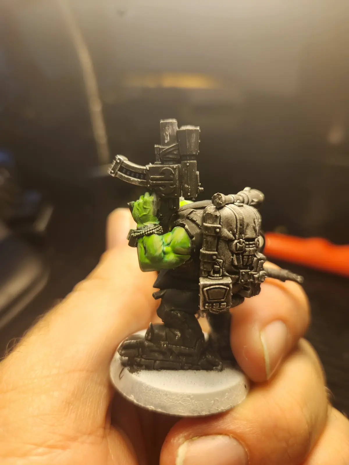 A hand holds a painted miniature of a green-skinned, muscular orc-like warrior from the Warhammer 40,000 universe, wearing…