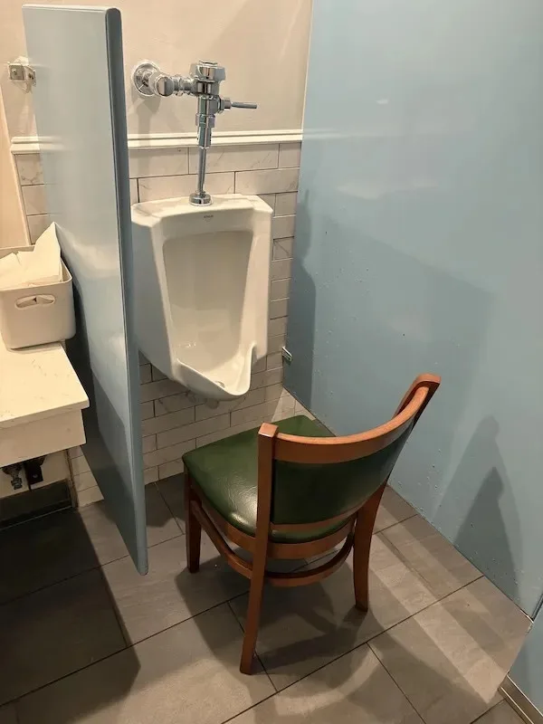 A photo of a public restroom stall with a white urinal mounted on a tiled wall, flanked by light blue privacy partitions. In…