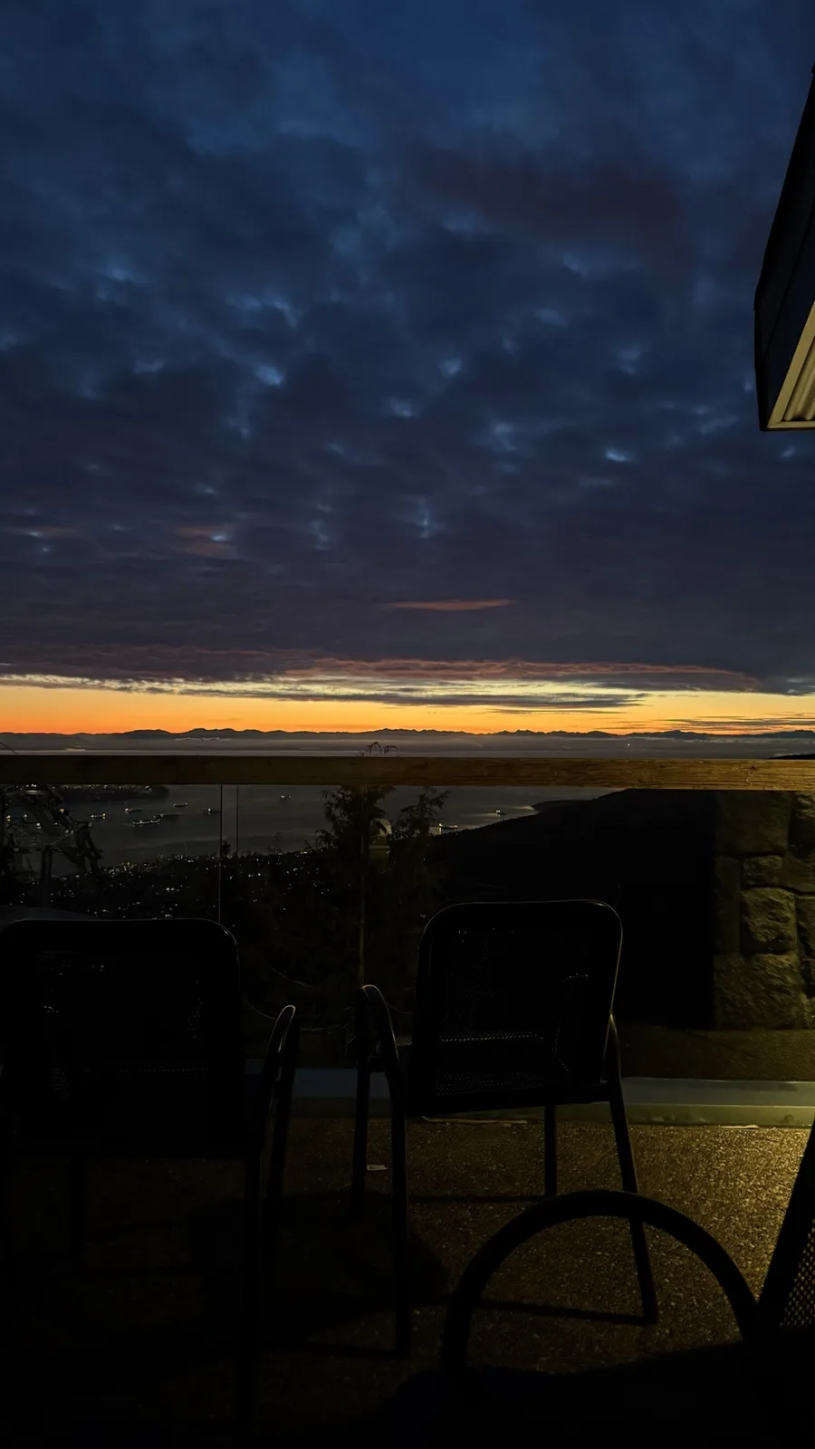 A dimly lit balcony at dusk shows two empty black mesh chairs facing a scenic view of a cityscape and water under a dramatic…