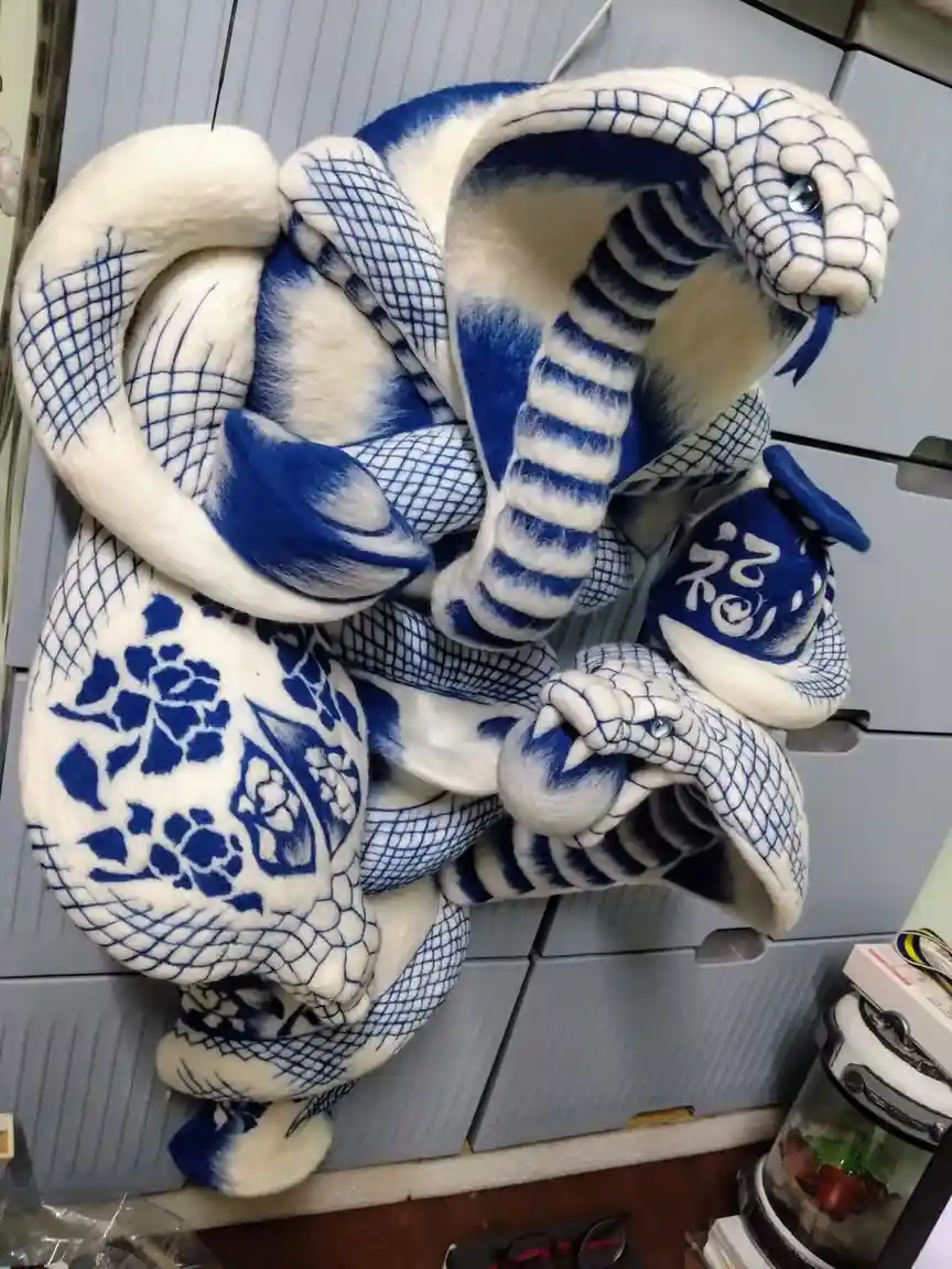 A large plush cobra toy with blue and white porcelain-style patterns, coiled and holding a small blue and white porcelain…