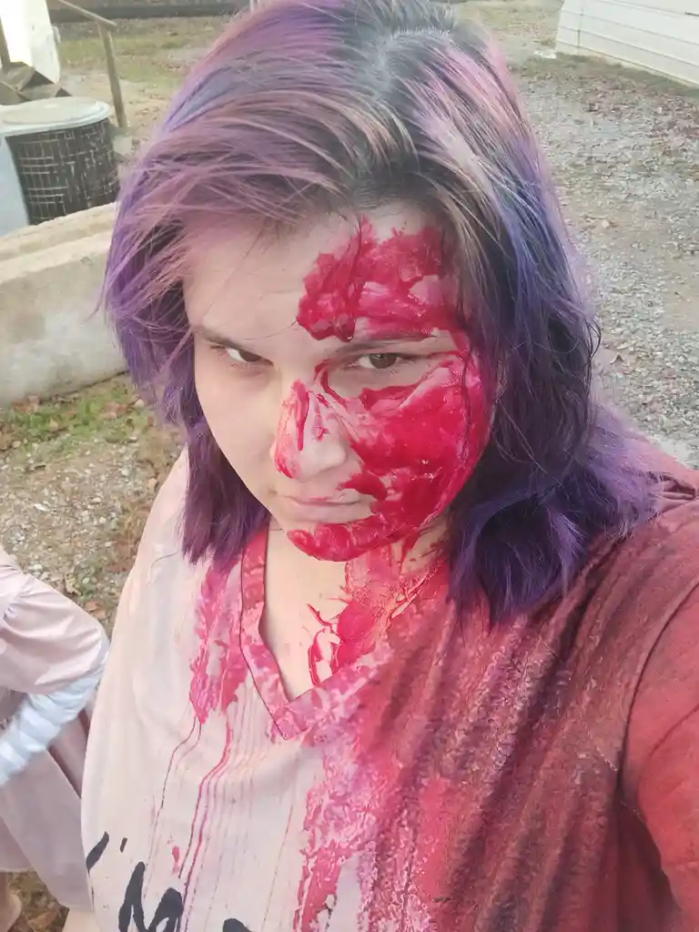 A person with purple hair has bright red paint smeared across half their face and neck, resembling blood, while wearing a…