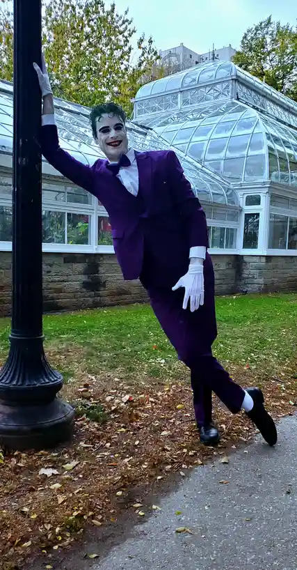 A person dressed as the Joker from Batman, wearing a purple suit, white gloves, and makeup with a wide grin, leans against a…