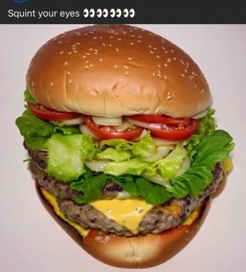 A close-up photograph of a large, appetizing cheeseburger with sesame seed buns, lettuce, tomato, onion, and a beef patty…