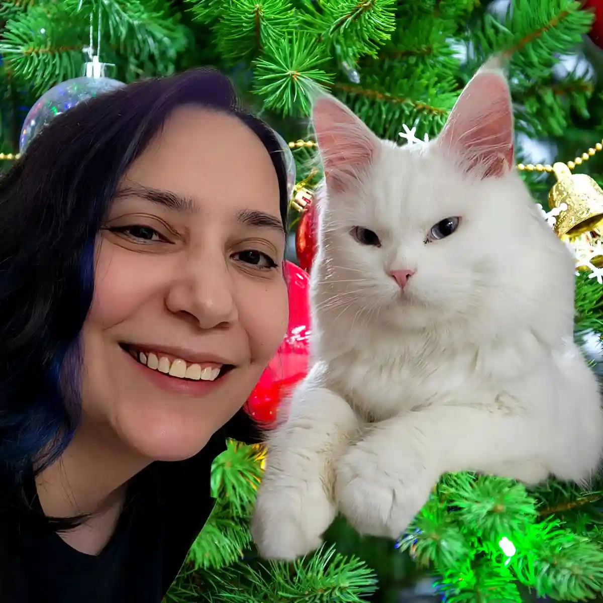 A selfie shows a smiling woman with dark hair and blue highlights next to a fluffy white cat perched on a decorated green…