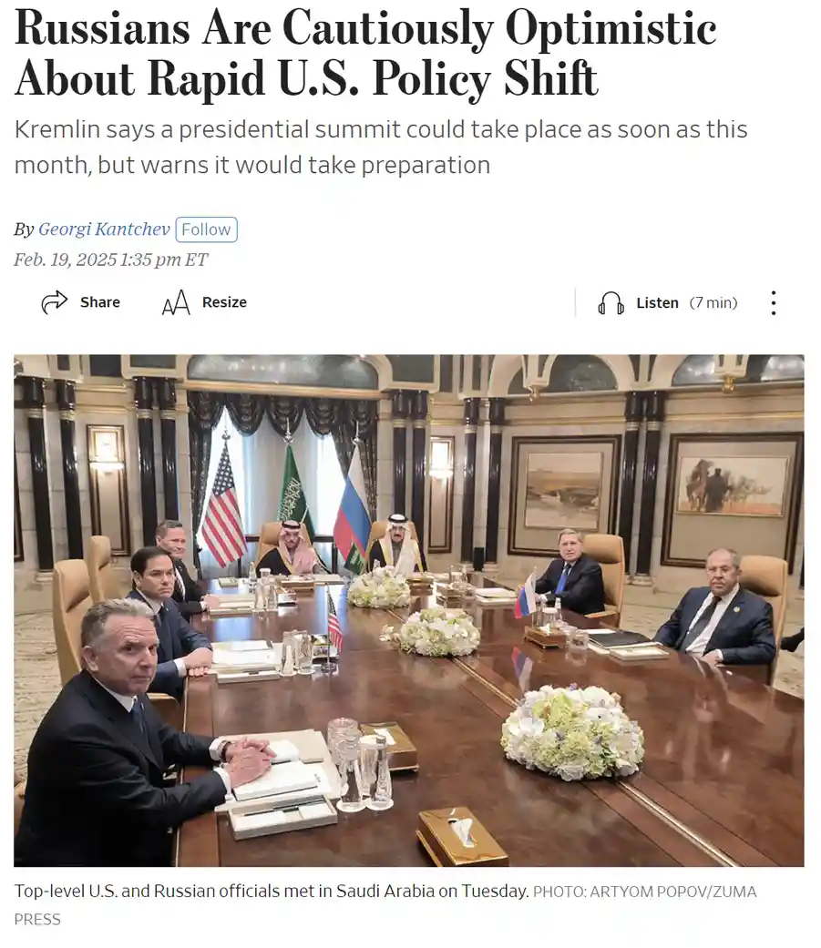 A formal diplomatic meeting photo shows several men in suits seated around a long wooden table with American, Russian, and…