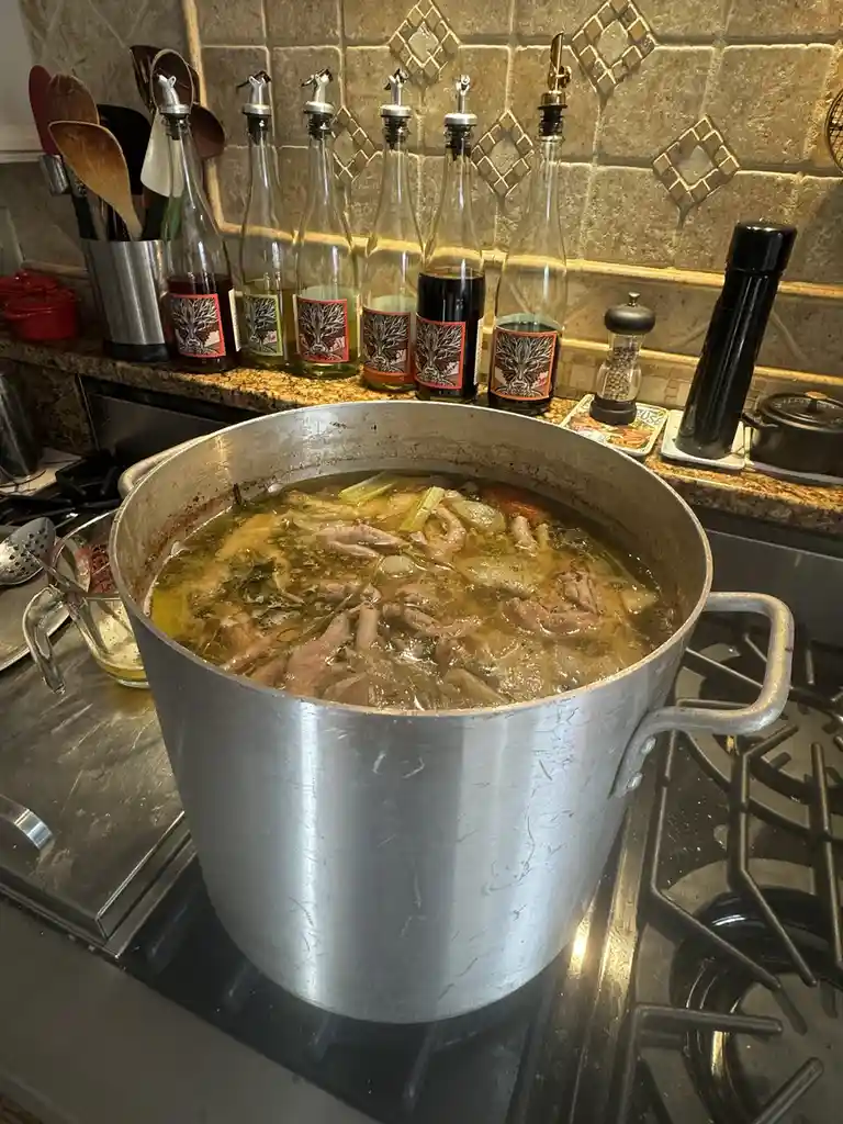 A large, silver stockpot sits on a gas stove, filled with a simmering, murky broth containing visible pieces of meat and…