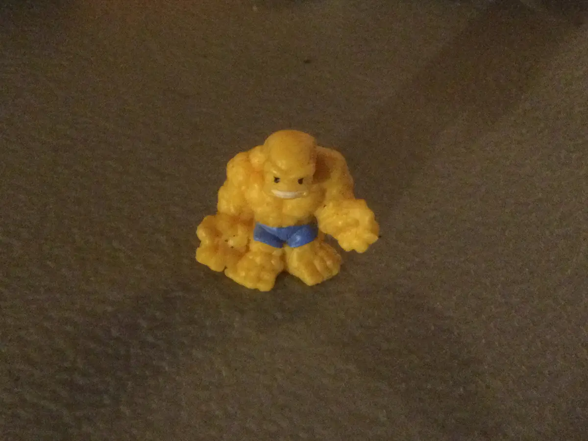 A small, yellow, plastic toy figure resembling a muscular humanoid with a stern expression and blue shorts sits on a dark…