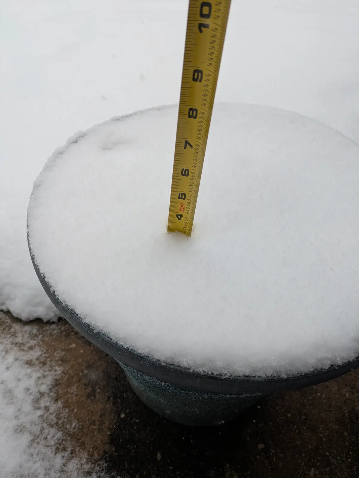 A yellow measuring tape is stuck vertically into a thick layer of snow piled on a dark, round outdoor object, showing the…