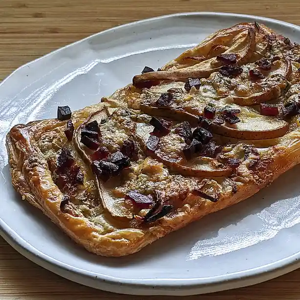 A close-up photo shows a golden-brown rectangular tart on a white ceramic plate, resting on a wooden surface. The tart is…