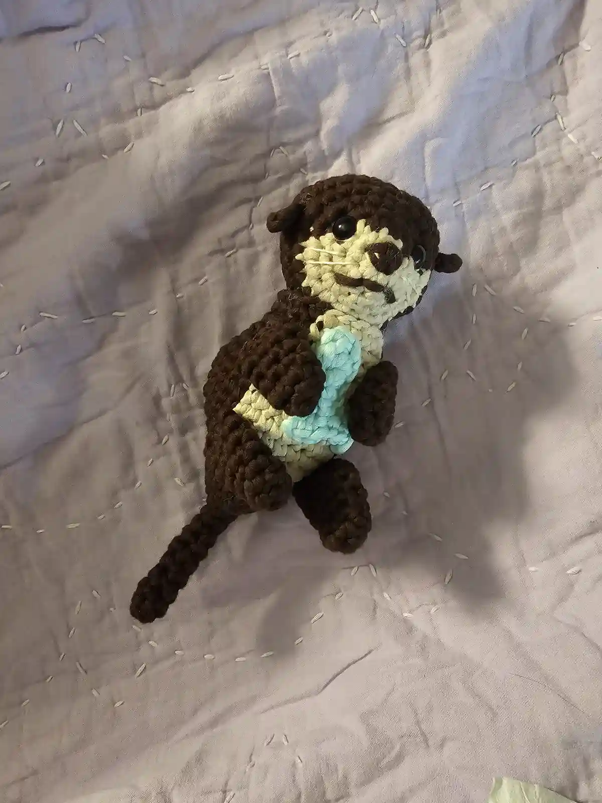 A handmade crocheted otter toy with brown and cream-colored fur, black button eyes, and a light blue crocheted baby otter…
