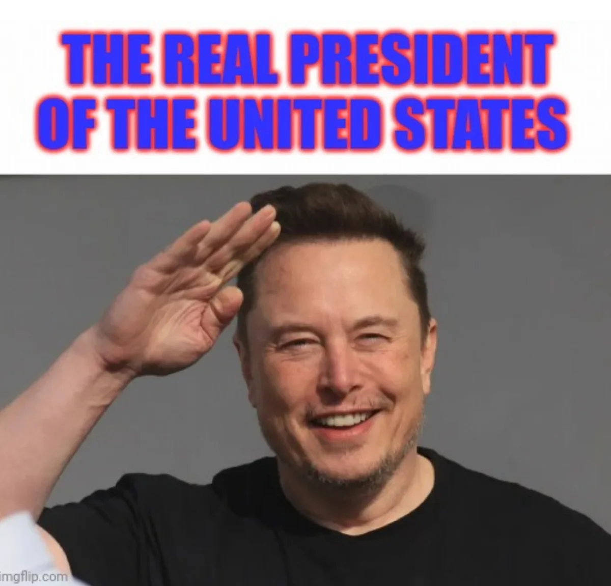 A man with short dark hair and a beard, wearing a black t-shirt, smiles broadly while raising his right hand in a salute…
