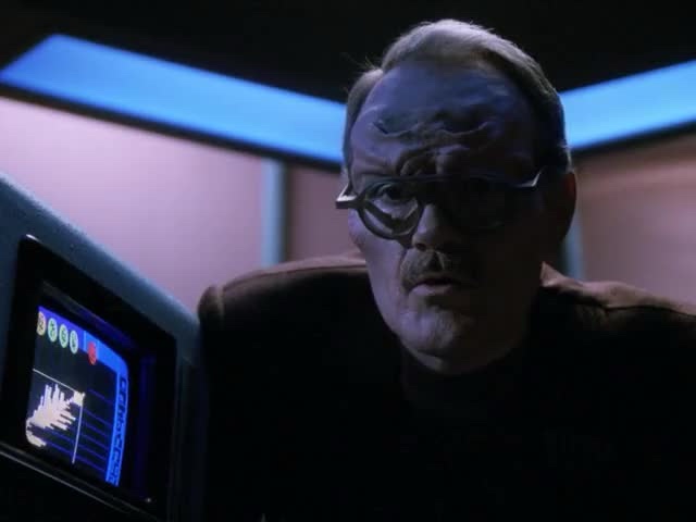 A blue-skinned, bespectacled alien man in a dark uniform stares intently at a glowing blue screen displaying a graph and…