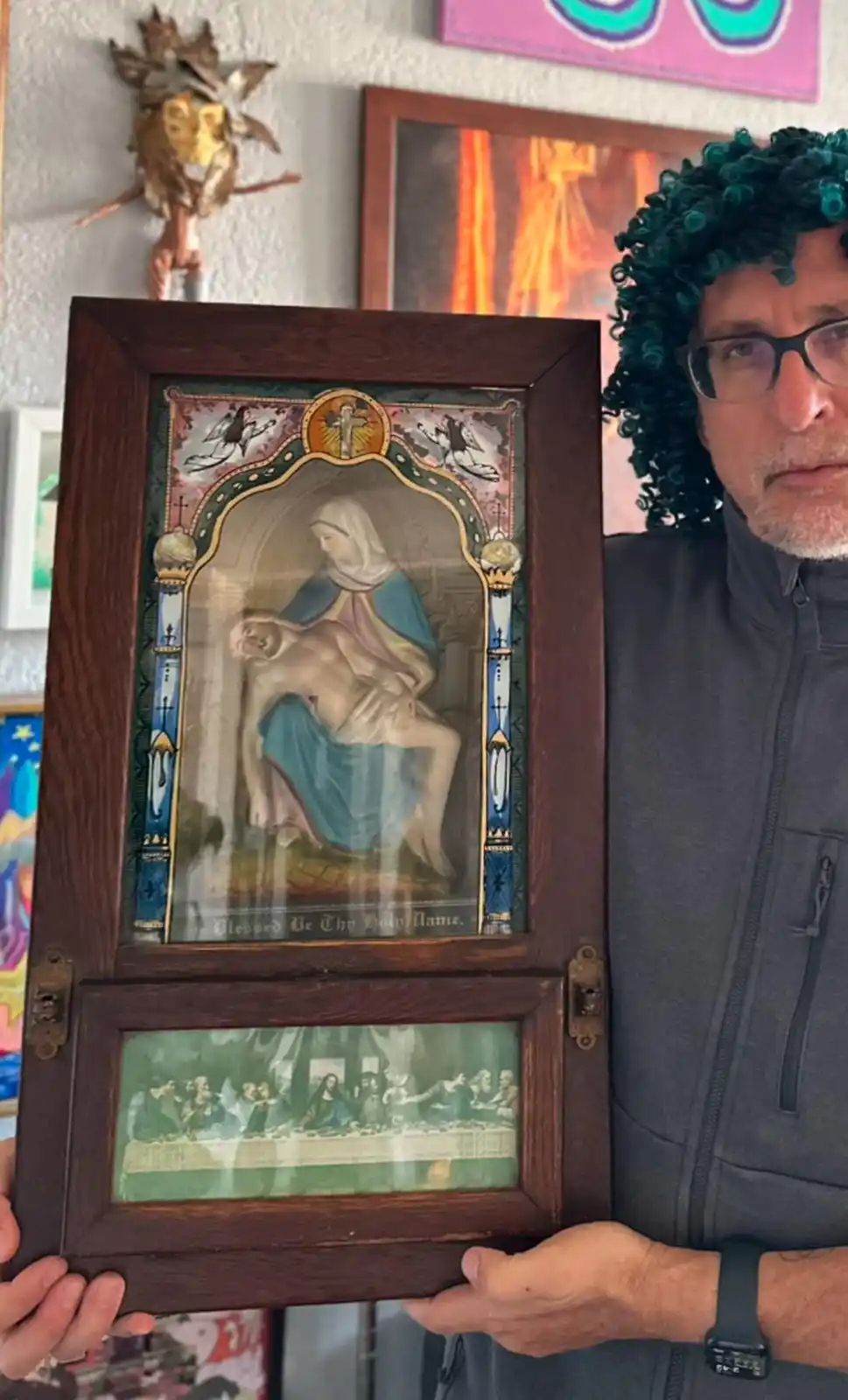 A man wearing glasses and a green curly wig holds a wooden-framed religious artifact. The top panel depicts the Pietà (Mary…