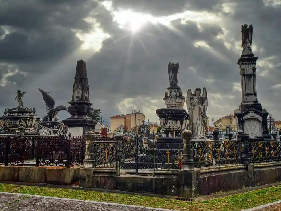 A dramatic cemetery scene with ornate gravestones and angel statues under a cloudy sky with bright sun rays breaking…