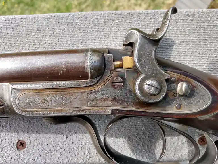 A close-up photo of an ornate, antique percussion pistol with intricate floral engravings on its metalwork and wooden grip…