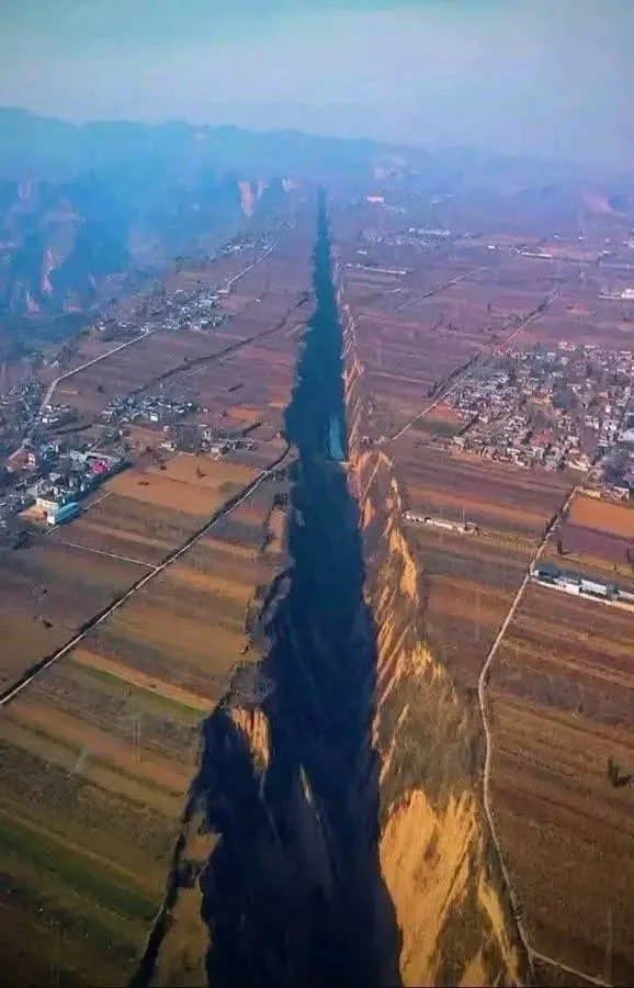 An aerial view shows a massive, deep, and long fissure cutting through a rural landscape with fields and villages on either…