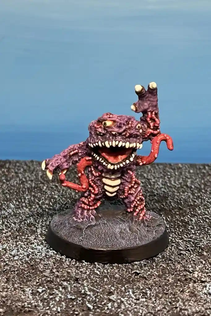 A painted miniature monster figurine with purple and red textured skin, multiple eyes, sharp teeth, and tentacle-like limbs…