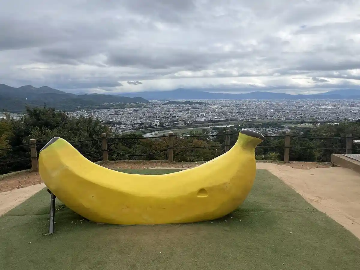 A giant yellow banana-shaped bench sits on artificial grass overlooking a sprawling city and mountains under a cloudy sky…