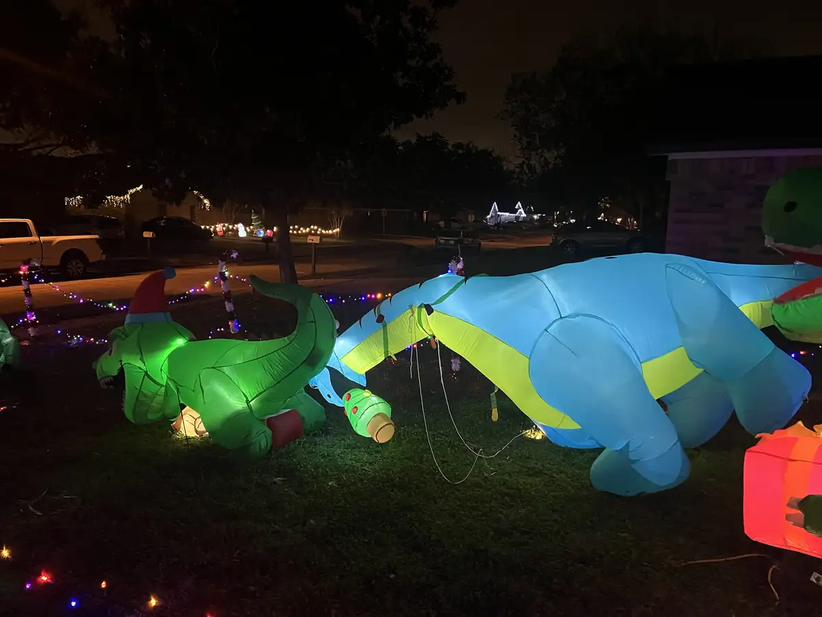 At night, two large inflatable dinosaurs—one green wearing a red Santa hat, the other blue with a yellow stripe—are lying on…