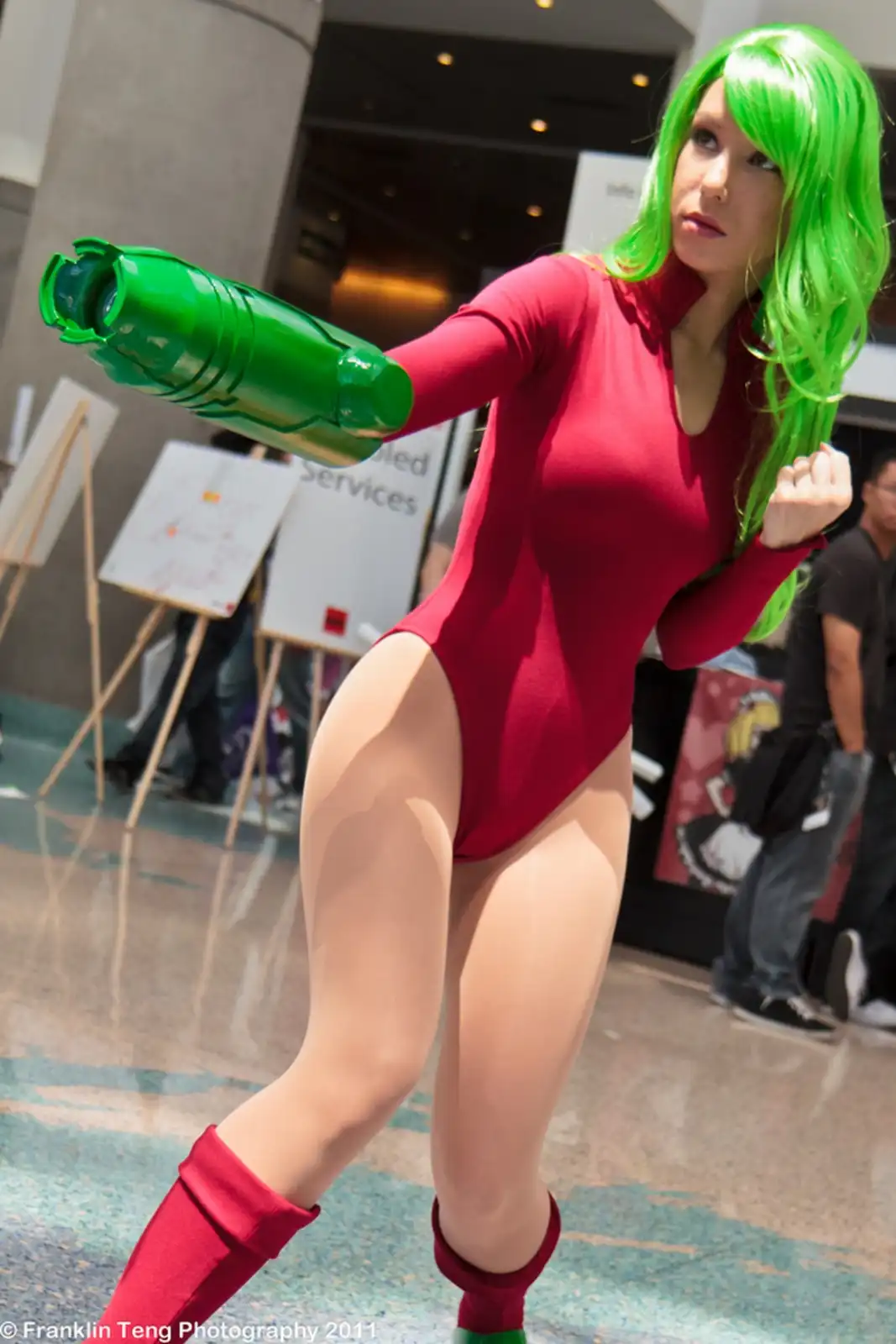 A cosplayer with bright green hair and a red bodysuit poses in a dynamic stance, holding a large green lantern-like weapon…