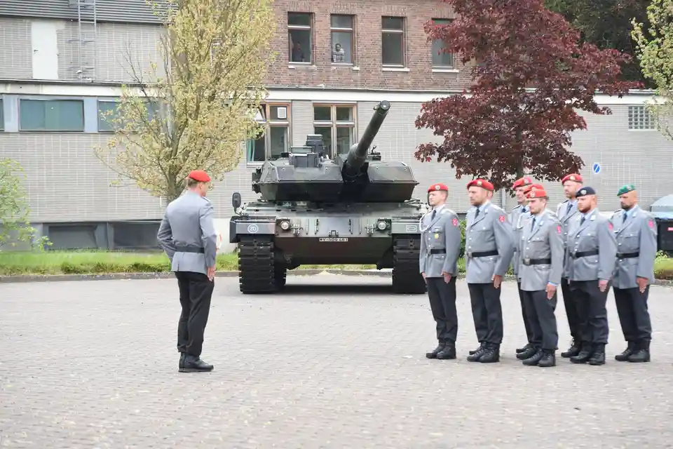 A group of soldiers in grey uniforms and red berets stand at attention in front of a military tank. One soldier faces the…