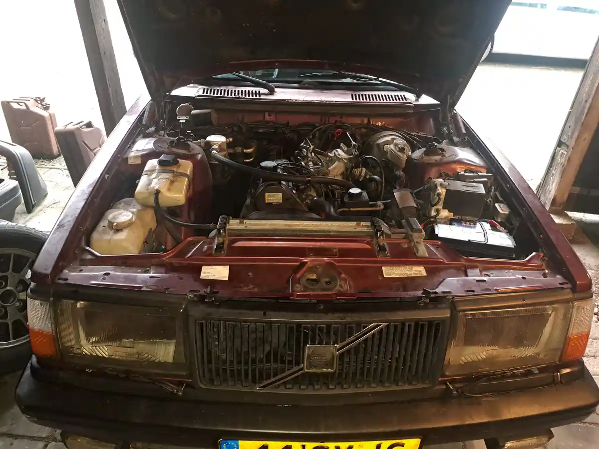 A front-facing photo of a maroon Volvo car with its hood open, revealing a visibly dirty and cluttered engine bay filled…