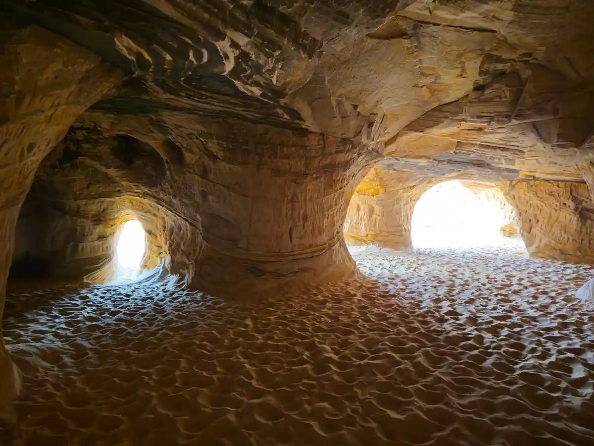 The image shows a natural rock formation with two arched openings letting in bright sunlight, illuminating rippled sand on…