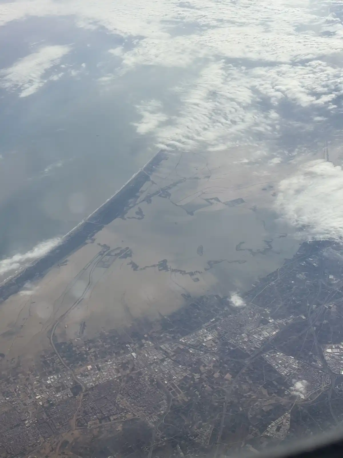 An aerial view from an airplane window shows a coastline with a large body of water, a sandy beach, and a developed urban…