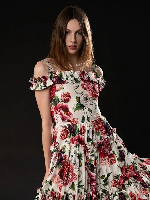 A woman with shoulder-length brown hair stands against a black background wearing a white dress covered in large red and…