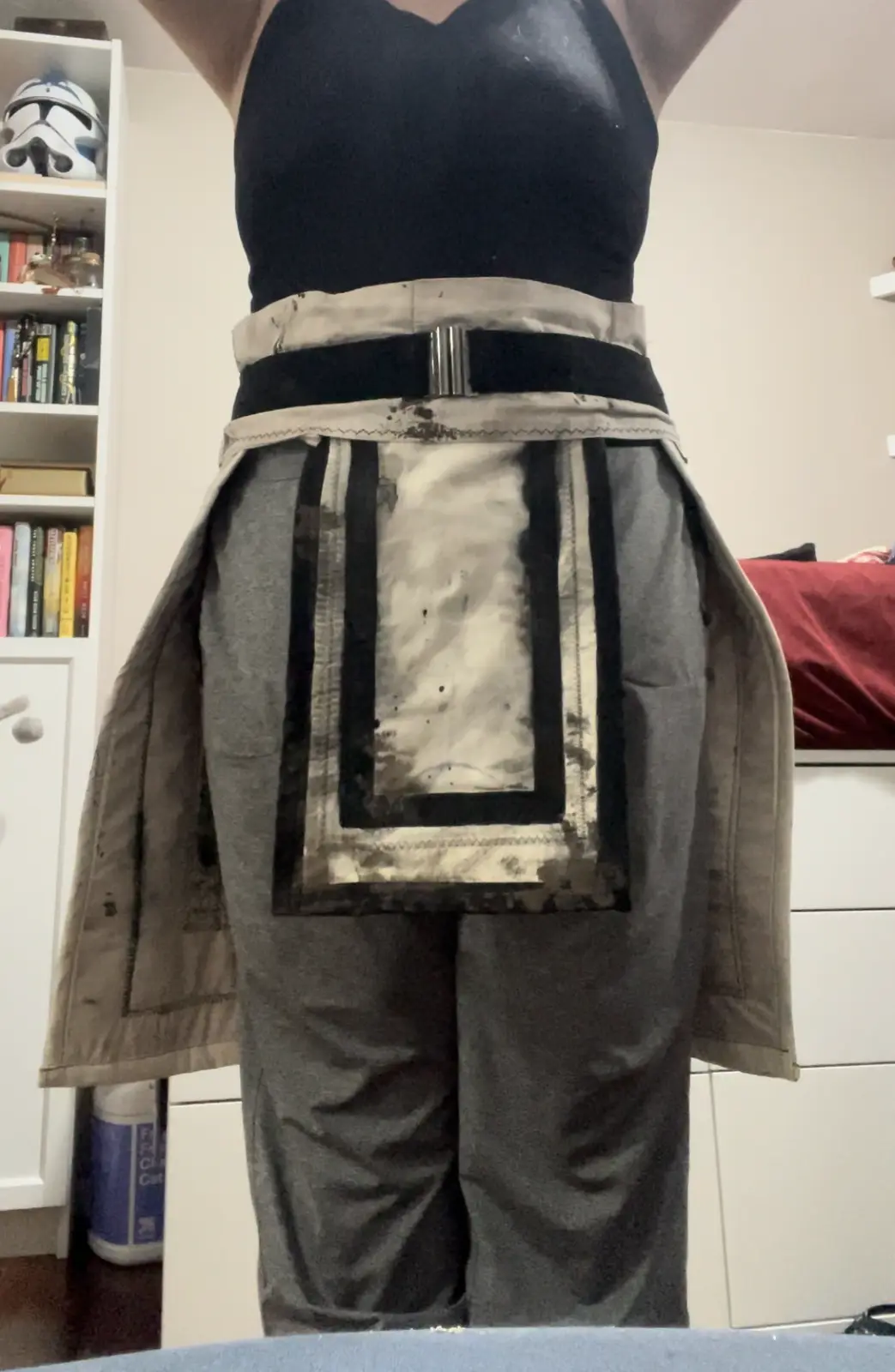 A person wearing a heavily stained grey apron with black trim and a silver buckle, standing with arms raised. The apron's…