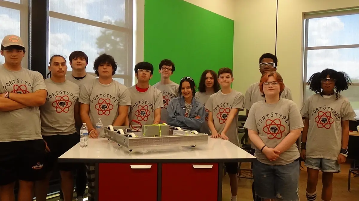 A group of teenagers wearing matching gray 'PORTOTYPES' t-shirts with a red atom logo stand around a table with a robot…