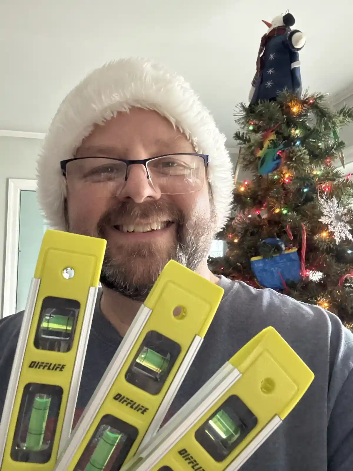 A man wearing a white fuzzy Santa hat and glasses smiles while holding three yellow spirit levels branded 'DIFFLIFE' in…