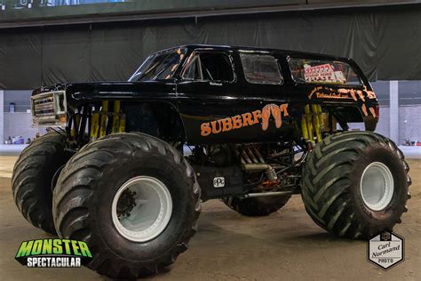 A black monster truck with oversized tires and exposed suspension is shown indoors. The word 'SUBERPOW' is written in orange…