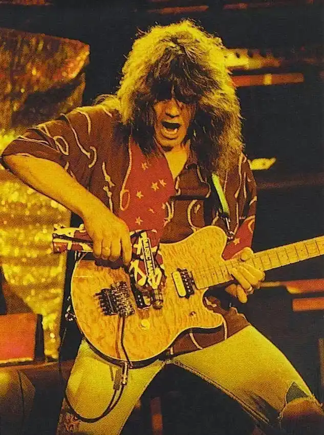 A man with long, shaggy hair and a patterned shirt is intensely playing an electric guitar on stage under warm stage…