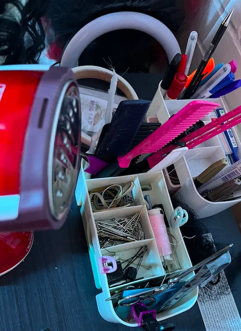 A cluttered desk with a white organizer holding pens, combs, and scissors, alongside a red electric razor and a container of…