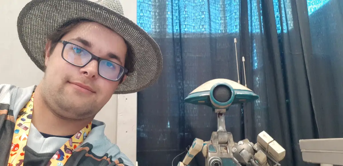 A person wearing glasses and a mesh hat poses beside a robot prop, both looking toward the camera. The robot has a…
